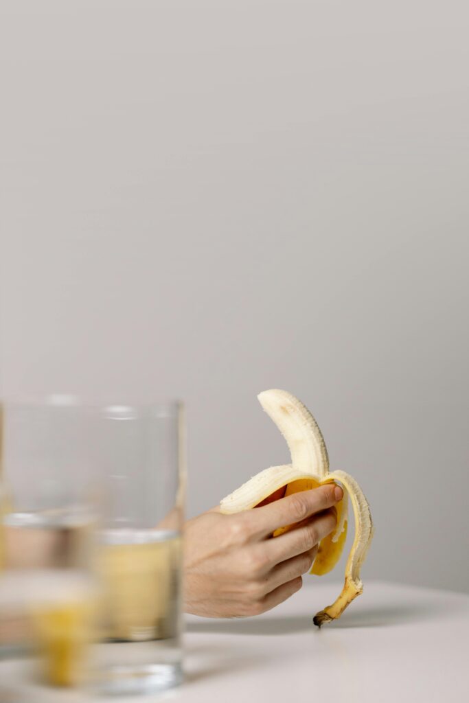 A minimalist photo of a hand holding a peeled banana next to a glass of water on a white surface.